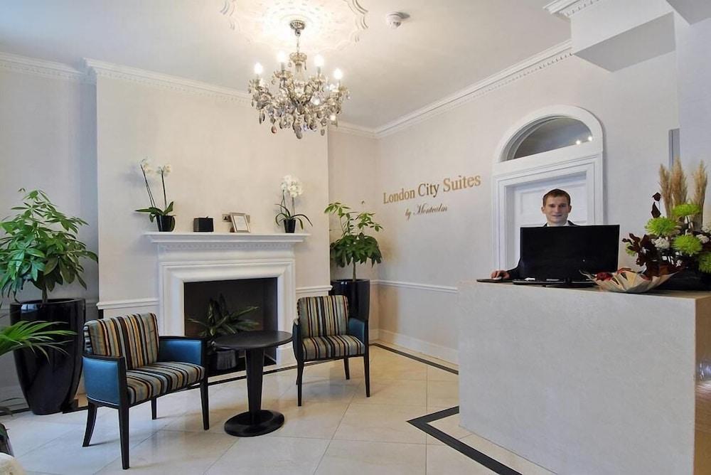 Montcalm Brewery Townhouse Hotel London Exterior photo The photo shows the interior of a reception area in a hotel called "London City Suites." The space features a light-colored wall with the hotel's name displayed. There is a stylish chandelier hanging from the ceiling, providing warm lighting. 

In th