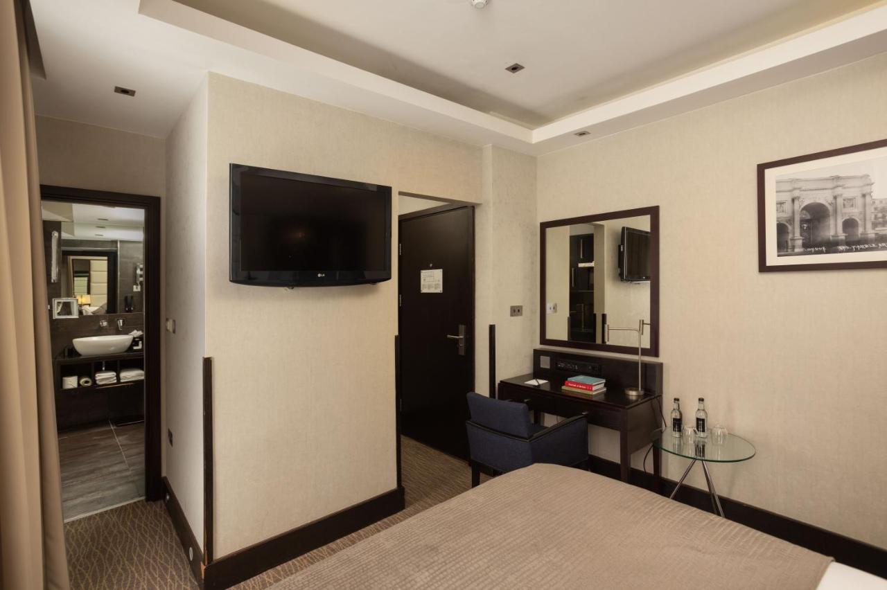 Montcalm Brewery Townhouse Hotel London Exterior photo The photo shows a hotel room featuring a double bed with a neutral-colored bedspread. On one wall, there is a flat-screen TV mounted. Adjacent to the bed, there is a stylish desk with a chair and a small glass table. A mirror is positioned above the 