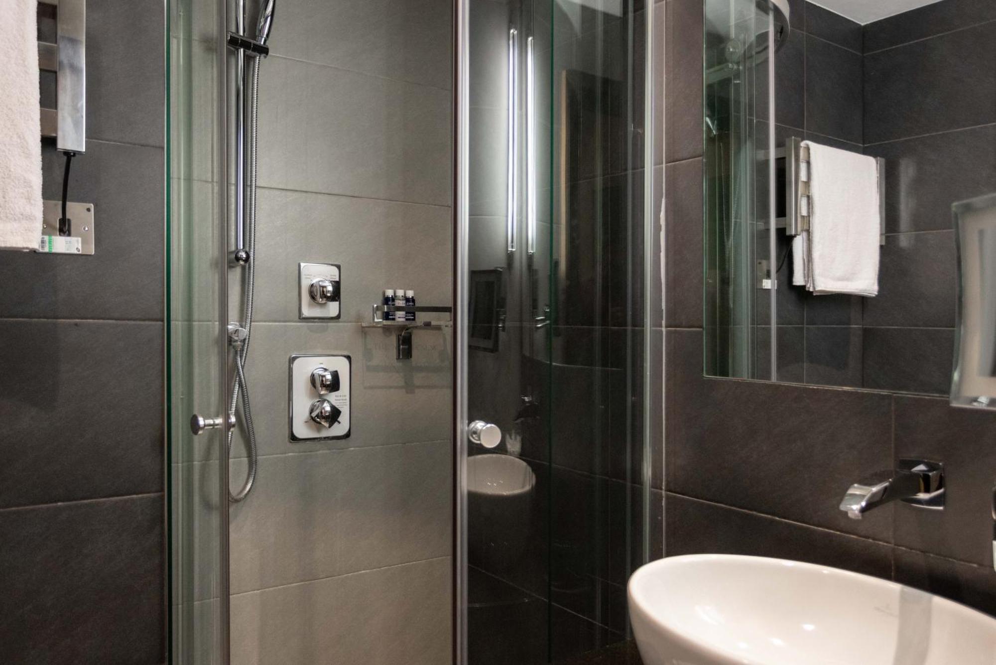 Montcalm Brewery Townhouse Hotel London Exterior photo The photo shows a modern bathroom with a glass shower enclosure. The shower features a handheld showerhead and various controls mounted on the wall. The walls are tiled in dark, sleek tones, contributing to a contemporary aesthetic. There is a round 