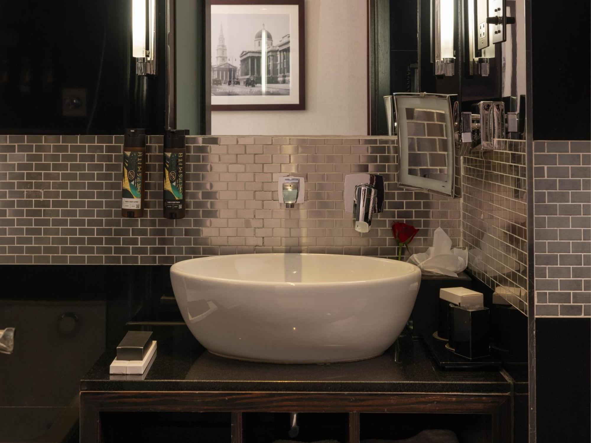 Montcalm Brewery Townhouse Hotel London Exterior photo The photo shows a modern bathroom with a stylish design. It features a large, round white sink placed on a dark wooden vanity. The walls are adorned with a mix of gray and silver tile, creating a sleek and contemporary look. Above the sink, there is 