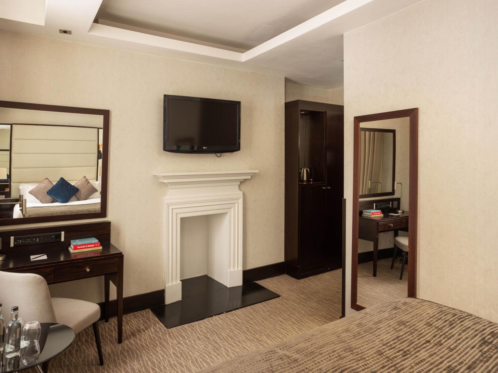 Montcalm Brewery Townhouse Hotel London Exterior photo The photo depicts a stylish hotel room. It features a double bed, a large flat-screen TV mounted on the wall, and a decorative fireplace. There is a wooden desk with a chair beside it, as well as a mirror hanging on the wall. The decor is modern and 