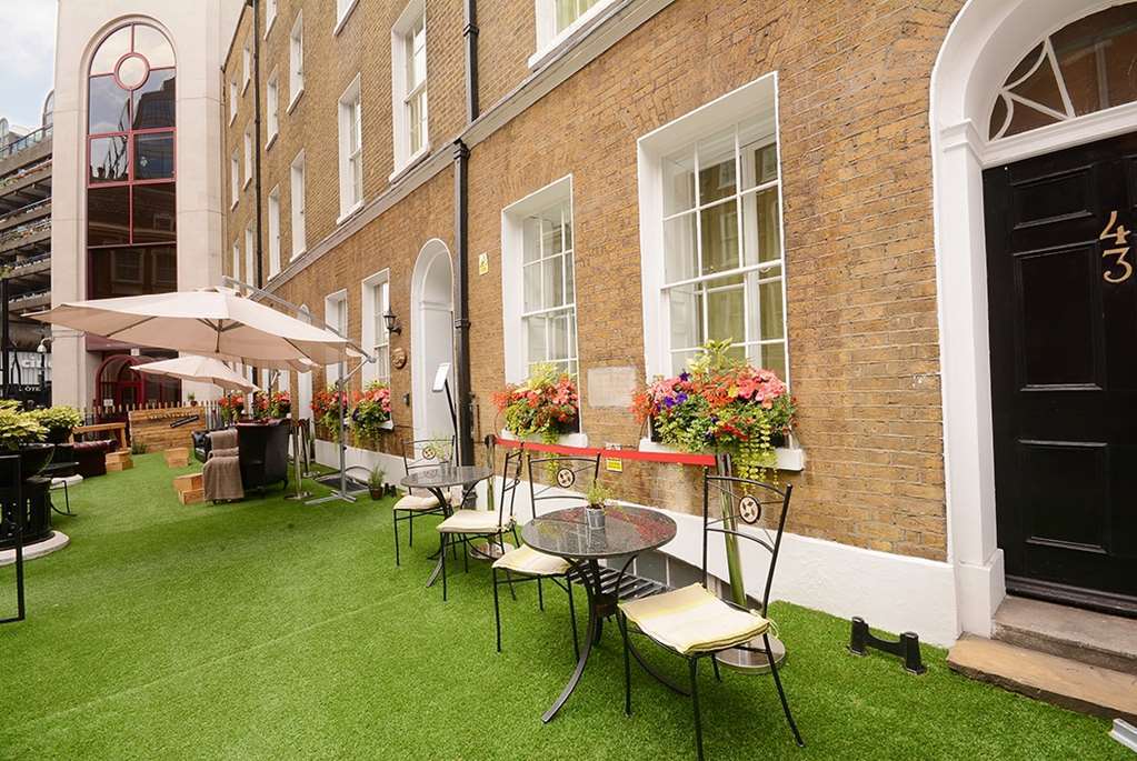 Montcalm Brewery Townhouse Hotel London Exterior photo The photo depicts a charming outdoor area in front of a building with a warm, textured brick facade. There is a neatly maintained lawn, likely made of artificial grass, and several small round tables with chairs are arranged outside. Flower pots with