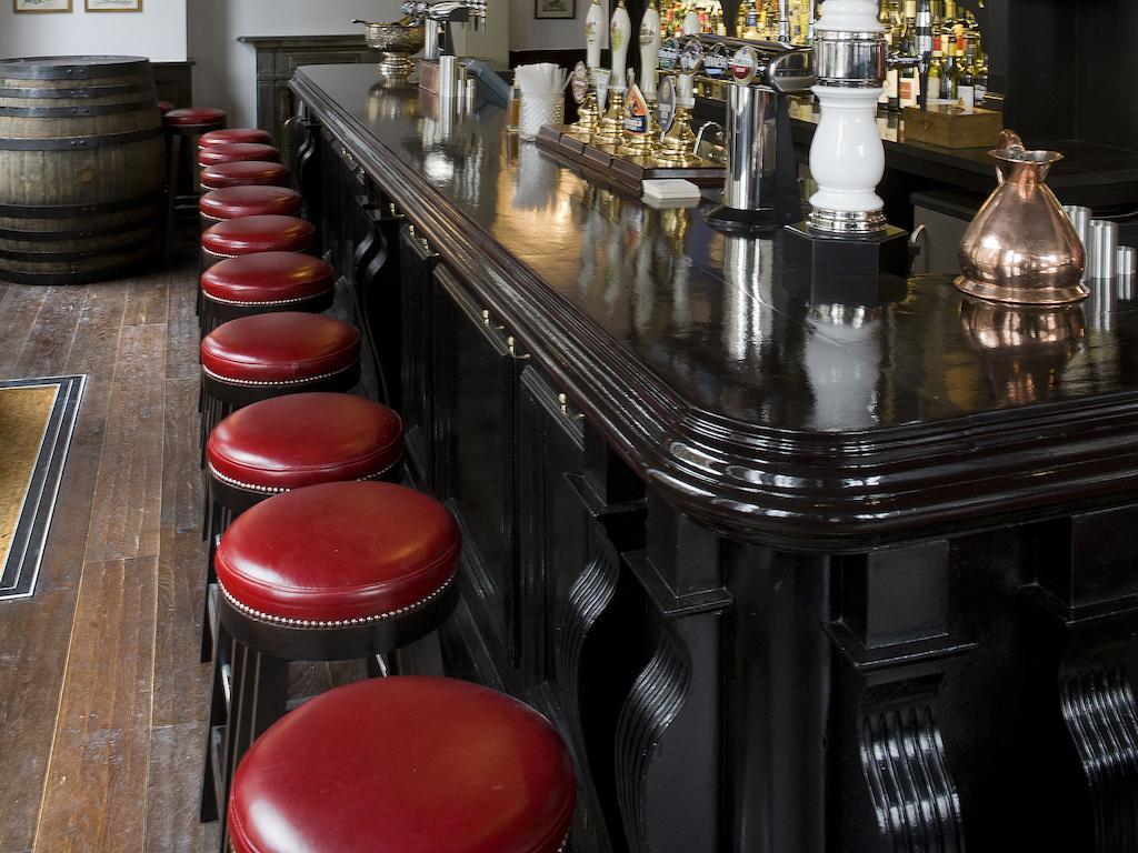 Montcalm Brewery Townhouse Hotel London Exterior photo The photo shows a bar setting with a long, polished dark wooden bar counter. Attached to the counter are several bar stools with red leather seats. Behind the bar, there are various bottles, suggesting a well-stocked bar. The flooring appears to be w