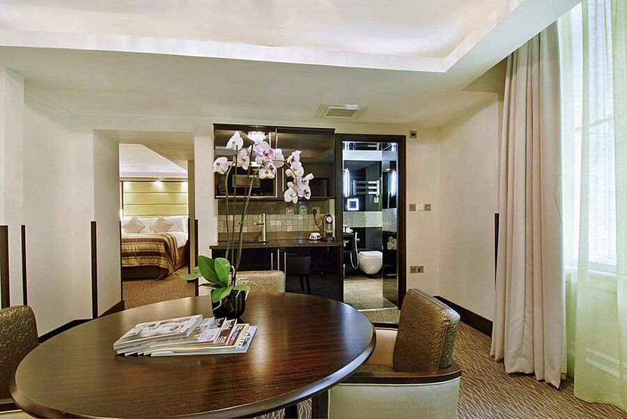 Montcalm Brewery Townhouse Hotel London Exterior photo The photo shows a stylish hotel room or suite. In the foreground, there is a round dining table with a few magazines on it. Surrounding the table are upholstered chairs. To the right, there are large windows with light-colored curtains, allowing natu