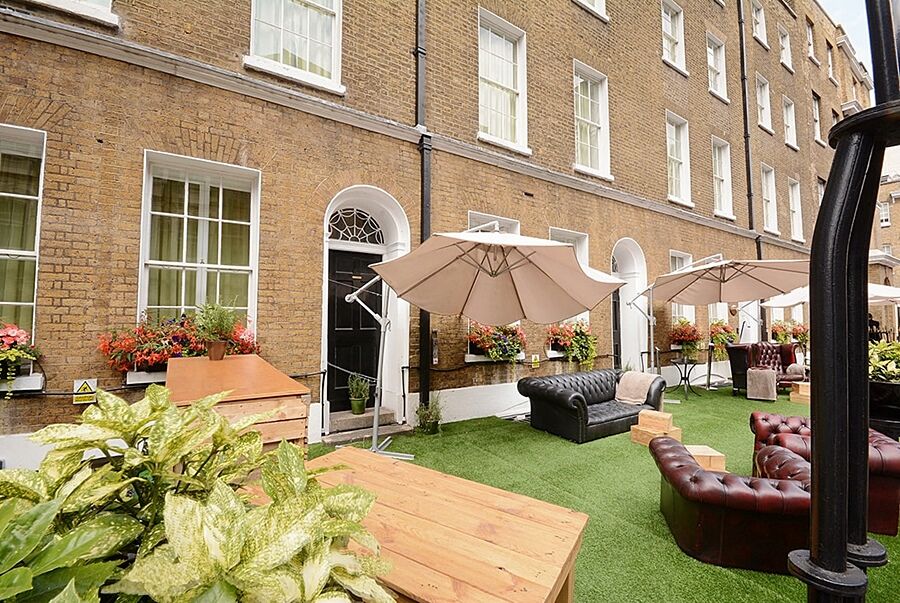 Montcalm Brewery Townhouse Hotel London Exterior photo The photo depicts an outdoor area of a building with a brick facade. There are several large windows visible, along with a wooden door. The setting features outdoor furniture, including couches in a dark leather finish and wooden tables. Large umbrel